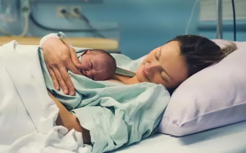 Eine Mutter hält ihr Neugeborenes im Arm, glücklich und erschöpft. Die Szene zeigt, wie lange nach Geburt im Krankenhaus die erste Bindung entsteht.