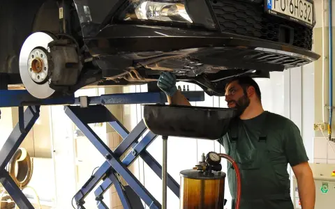Mechanik w warsztacie wymienia zużyty olej samochodowy. Podniesiony pojazd i naczynie na płyn wskazują na rutynową czynność serwisową.