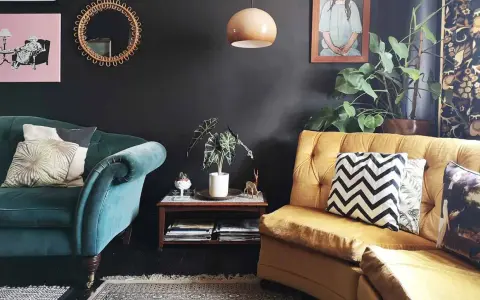 Habitación con poca luz, sofás de terciopelo verde y mostaza, cojines decorativos, plantas y arte en la pared.