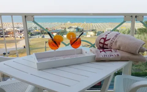 Due aperitivi arancioni e asciugamani con logo "Hotel Rivazzurra Rimini" sul tavolo, con vista sul rivazzurra mare e spiaggia attrezzata.