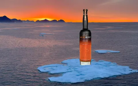 Eine Flasche New Riff Bourbon Whiskey steht auf einer Eisscholle im Meer. Der Sonnenuntergang taucht die Berge am Horizont in warmes Licht. Ein Moment der Ruhe für den besten Whisky der Welt.