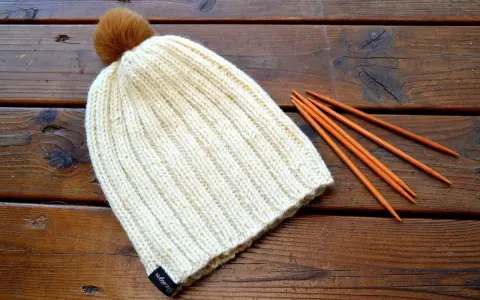 En ribbstickad mössa med en brun pompom ligger på ett träbord bredvid stickor. Perfekt för ett ribbstickad mössa gratis mönster.
