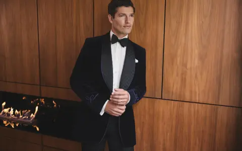 A man in a dark velvet tuxedo with a bow tie, embodying festive chic. He stands by a modern fireplace with flames.