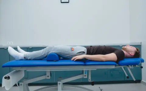 Mężczyzna leży na kozetce rehabilitacyjnej, z podparciem pod kolanami. Rozpoczęła się jego sis rehabilitacja.