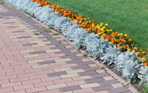 Un camino de adoquines de colores bordea unos parterres de flores anaranjadas y amarillas, con plantas plateadas y césped verde.