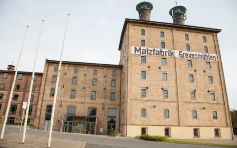 Die imposante Malzfabrik Grevesmühlen, ein Backsteingebäude mit markanten Türmen, steht im hellen Tageslicht.