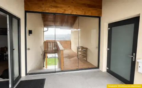 Moderner, verglaster Windfang aus Aluminium mit Holzbalken und Blick auf eine Holzterrasse.