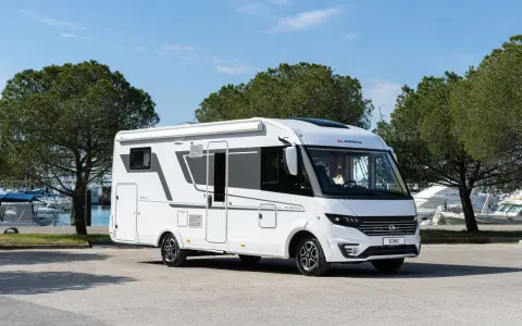 En vit Adria Sonic husbil, den lyxigaste husbilen, parkerad vid en marina med båtar och träd i bakgrunden.