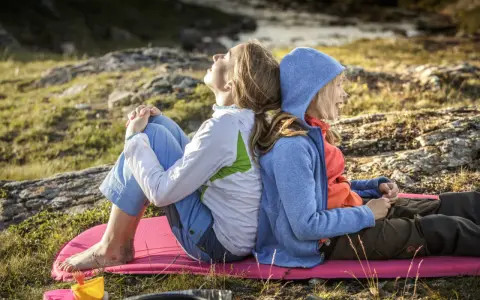 Två personer, en vuxen och ett barn, sitter rygg mot rygg på en liggunderlag i naturen. En härlig stund att vandra med barn.