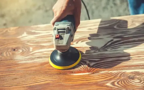 Main d'artisan utilisant une ponceuse pour enlever lasure sans poncer le bois. Le grain du bois est bien visible.