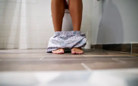 Nogi osoby siedzącej na toalecie, z opuszczonymi luźnymi spodniami. Czasem tak bywa, że występuje rzadkie oddawanie moczu.