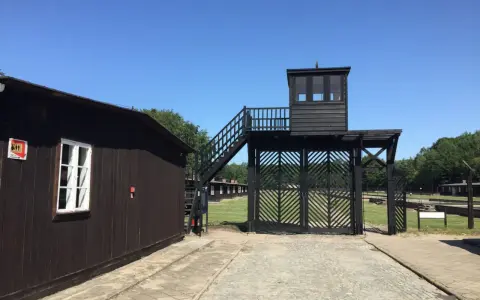 Brama i wieża strażnicza na terenie Muzeum Stutthof. Drewniane budynki i brukowany chodnik.