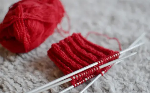 Rött pompom garn ligger bredvid ett stickat provstycke på stickor.