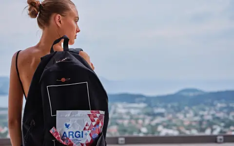 Młoda kobieta z plecakiem, w którym widać opakowanie ARGI+. W tle krajobraz miasta i wzgórz.