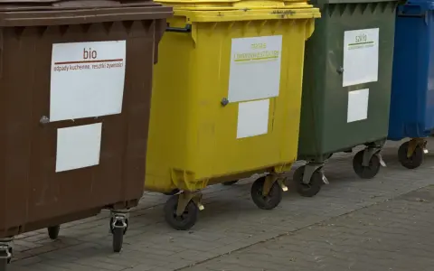 Rząd kolorowych pojemników na śmieci: brązowy (bio), żółty (metale i tworzywa sztuczne), zielony (szkło), niebieski (papier i tektura).