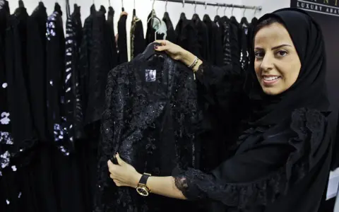 A woman smiles, holding a black lace abaya, showcasing elegant Saudi Arabia fashion.