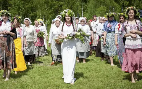 Grupa kobiet wiankach na głowach i tradycyjnych strojach idzie przez łąkę. Obraz nawiązuje do święta zielone świątki.