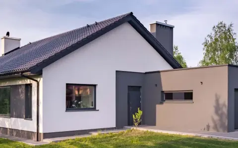 Nowoczesny dom z białymi i szarymi akcentami, prezentujący najmodniejsze kolory elewacji. Dachówka i komin w ciemnym odcieniu.