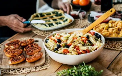 Pyszna sałatka makaronowa z oliwkami i pomidorkami, obok pieczywo i świeże oregano. Idealne na letni obiad, pokazujące co zrobić z oregano.