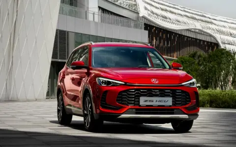 Czerwony MG ZS HEV, nowoczesny SUV hybrydowy, zaparkowany przed futurystycznym budynkiem.