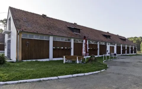 Długi budynek stajni z drewnianymi drzwiami i oknami, z parasolem i ławką przed nim.