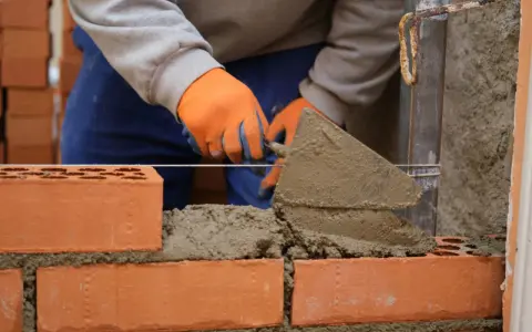 Budowa prostego grilla z cegły. Ręce w pomarańczowych rękawiczkach nakładają zaprawę kielnią na kolejne cegły.
