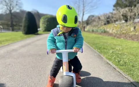 A child in a bright green helmet and teal jacket rides a woom wow balance bike on a sunny path.