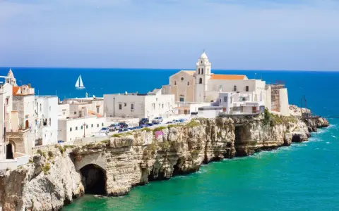 Un borgo bianco su scogliere a picco sul mare turchese, con una chiesa imponente. Un assaggio di cosa vedere in Puglia, con un veliero all'orizzonte.