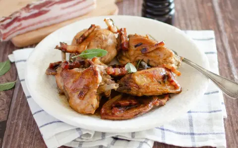 Delizioso coniglio in padella, dorato e succulento, guarnito con pancetta croccante e foglie di salvia fresca.