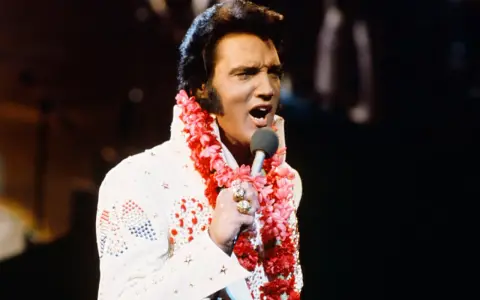 Elvis Presley singt in Hawaii, trägt einen weißen Anzug mit Blumenkette und Mikrofon.