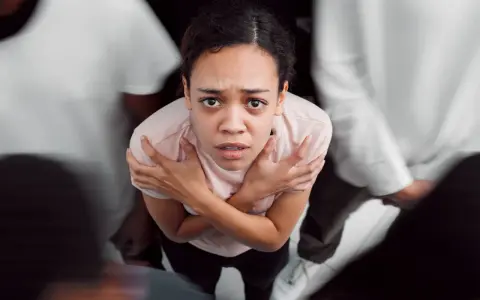 Donna spaventata, circondata da figure sfocate, con le braccia incrociate sul petto. Potrebbe rappresentare i disturbi psicosomatici, un elenco di sintomi fisici legati allo stress.