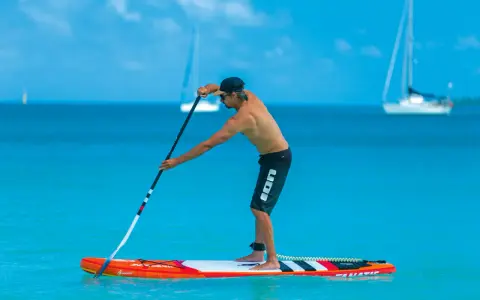Mann stellt sein SUP Paddel ein, während er auf einem Board im türkisfarbenen Wasser steht. Segelboote sind im Hintergrund zu sehen.