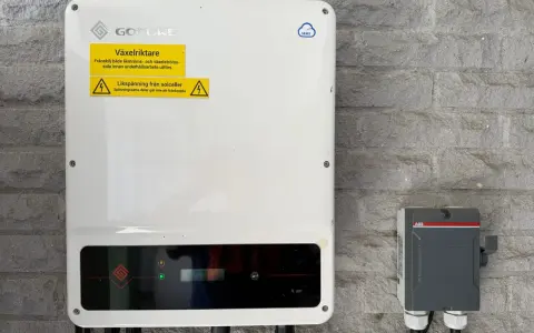 En vit GoodWe växelriktare 10kW monterad på en grå stenvägg. Bredvid sitter en grå ABB-box.