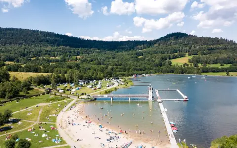 Słoneczny dzień nad zalewem w Starej Morawie. Ludzie odpoczywają na plaży i w wodzie, a w tle widać zielone wzgórza.