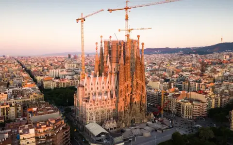 Widok z lotu ptaka na Sagrada Familia w Barcelonie, wciąż w budowie, z dźwigami górującymi nad miastem. Słońce oświetla fasadę, tworząc ciepłą poświatę, jakby przygotowując się do wieczornych mszy.