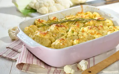 Pyszna keto zapiekanka z kalafiora w różowej naczyniu, udekorowana rozmarynem. Obok świeże różyczki kalafiora.