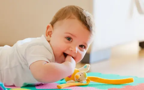 Uśmiechnięte 11 miesięczne dziecko leży na macie, gryząc paluszek. Obok pluszowa zabawka.
