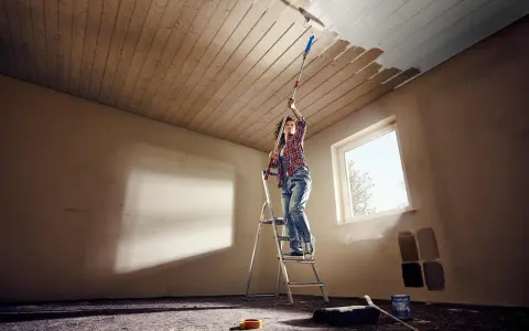 Frau streicht mit einer Teleskopstange und Rolle die Holzpaneele an der Decke weiß.