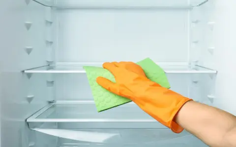Main d'une personne portant un gant orange, essuyant une étagère de réfrigérateur vide avec une éponge verte. Le nettoyage du frigo est terminé.