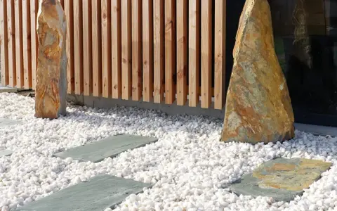 Deux pierres dressées sur une allée en gravier blanc, bordée de dalles grises et d'une palissade en bois.
