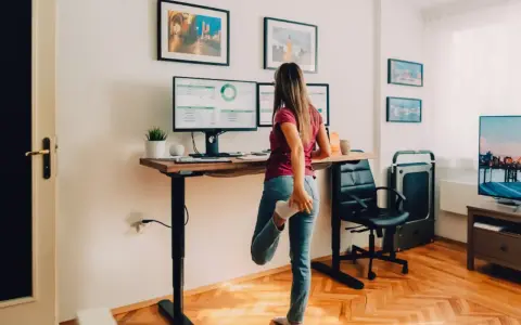 Femme s'étirant près de son bureau réglable en hauteur, pour un confort au bureau optimal. Deux écrans affichent des graphiques.