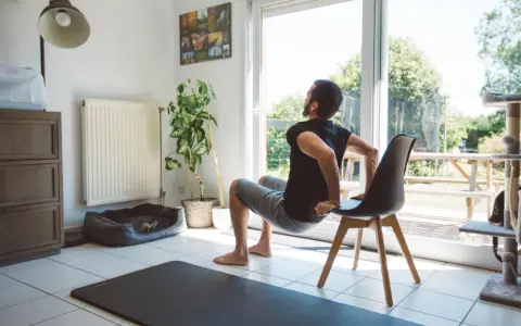 Un homme fait des dips sur une chaise, une idée de quel sport faire à la maison. Un tapis de yoga est au sol.