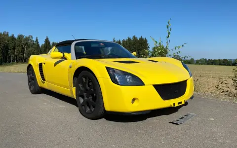 Żółty Opel Speedster na tle lasu i pól. Słońce pięknie podkreśla jego sportową sylwetkę.