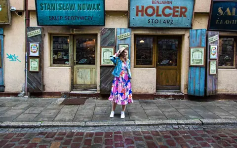 Kobieta w kolorowej sukience i jeansowej kurtce stoi przed sklepami w Krakowie. W tle szyldy "Stanisław Nowak Sklep Spożywczy" i "Benjamin Holcer Stolaz".