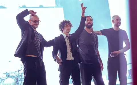 Matty Healy i zespół The 1975 na scenie. Muzycy pozdrawiają publiczność, jeden z nich unosi rękę w geście pokoju.