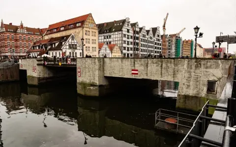 Kamienny most nad ciemną wodą, z rzędem zabytkowych kamienic w tle. W oddali widać dźwigi budowlane.