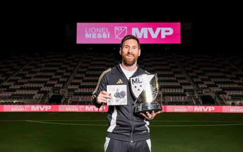 Lionel Messi sonríe en un estadio, sosteniendo un trofeo MLS MVP y un libro. El letrero del estadio dice "LIONEL MESSI MVP".