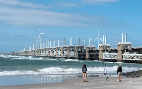 Dwie osoby stoją na plaży, obserwując falujące morze i imponującą zaporę z turbinami wiatrowymi. Takie morze jest w Holandii, chronione przez innowacyjne konstrukcje.