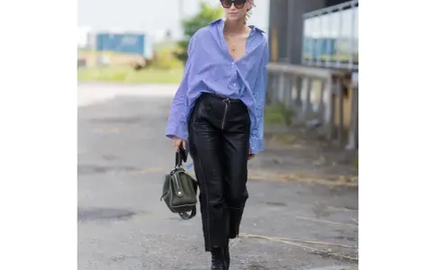 Donna con camicia a righe blu e bianche, occhiali da sole e un elegante outfit pantaloni ecopelle neri con zip frontale. Porta una borsa verde oliva.