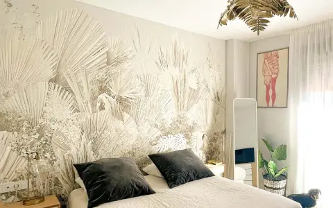Habitación con papel tapiz de palmeras, ideal para aprender cómo empapelar una pared. Cama con almohadas oscuras, espejo y planta.